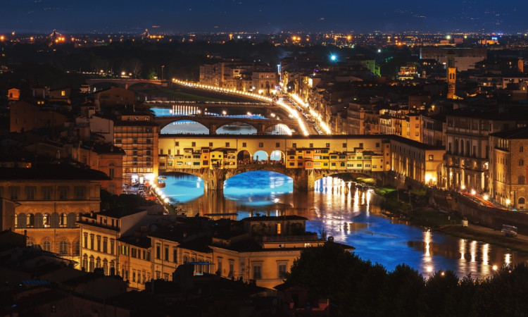  panoramic view of the city at night, with the famous bridge in florence visible from above. the river is flowing through it, and there's an illuminated bridge that connects to buildings on both sides. on one side of the photo, you can see tall buildings, while the other half shows greenery and trees. there are cars driving along the road near the water, adding movement to the scene.