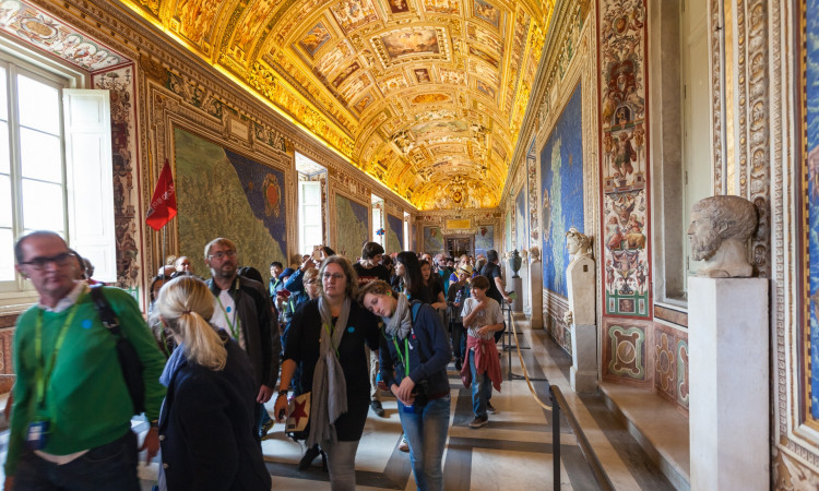 A group of tourists exploring the ornate hallway filled with colorful murals and intricate statues, with an intricately painted ceiling that includes various ancient figures and symbols, within the Sistine Chapel at the Vatican.